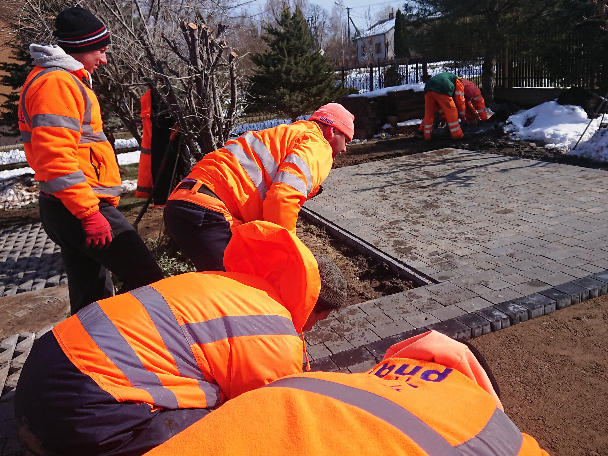Ekipa w pomarańczowych ubraniach roboczych układa szarą kostkę brukową na podsypce piaskowej, widoczne fragmenty śniegu i drzewa w tle.