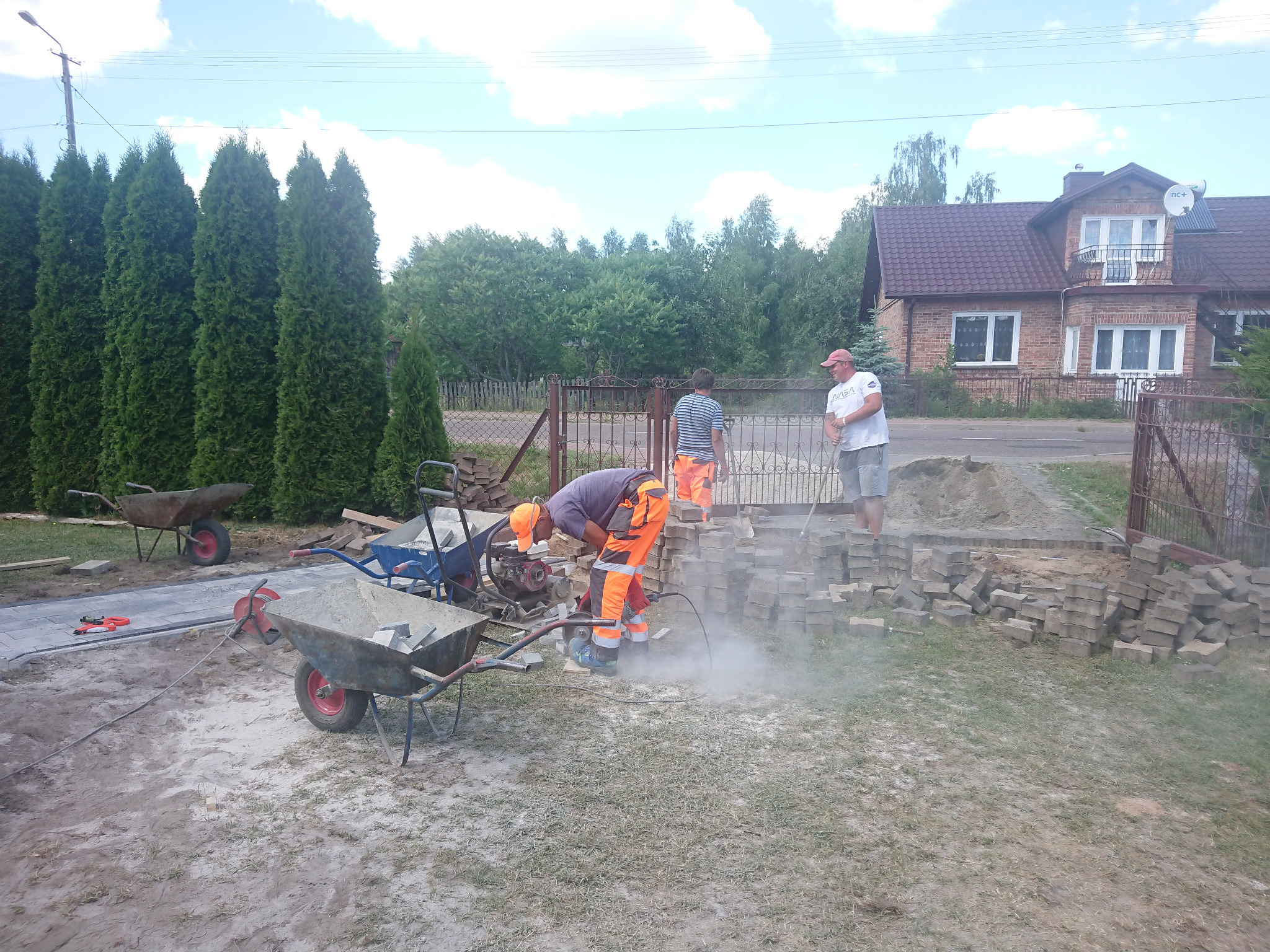 Pracownik w pomarańczowym kombinezonie tnie kostkę brukową szlifierką kątową, obok sterta kostki, taczki i częściowo ułożona nawierzchnia, w tle dom jednorodzinny i grupa osób.