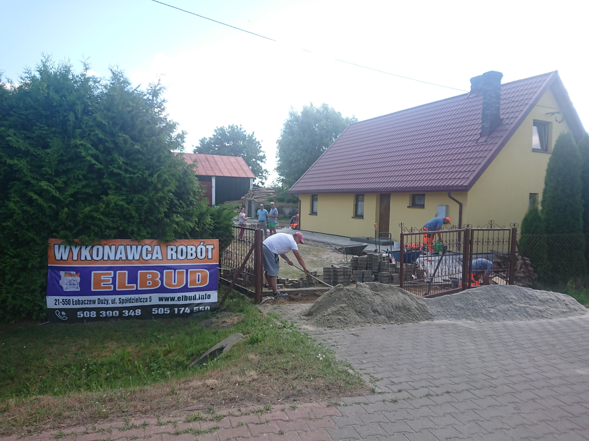 Pracownik w białej koszulce i szarych spodenkach wyrównuje podłoże pod kostkę brukową łopatą, widoczny stos kostki, piasek, dom jednorodzinny w tle i baner reklamowy firmy Elbud.