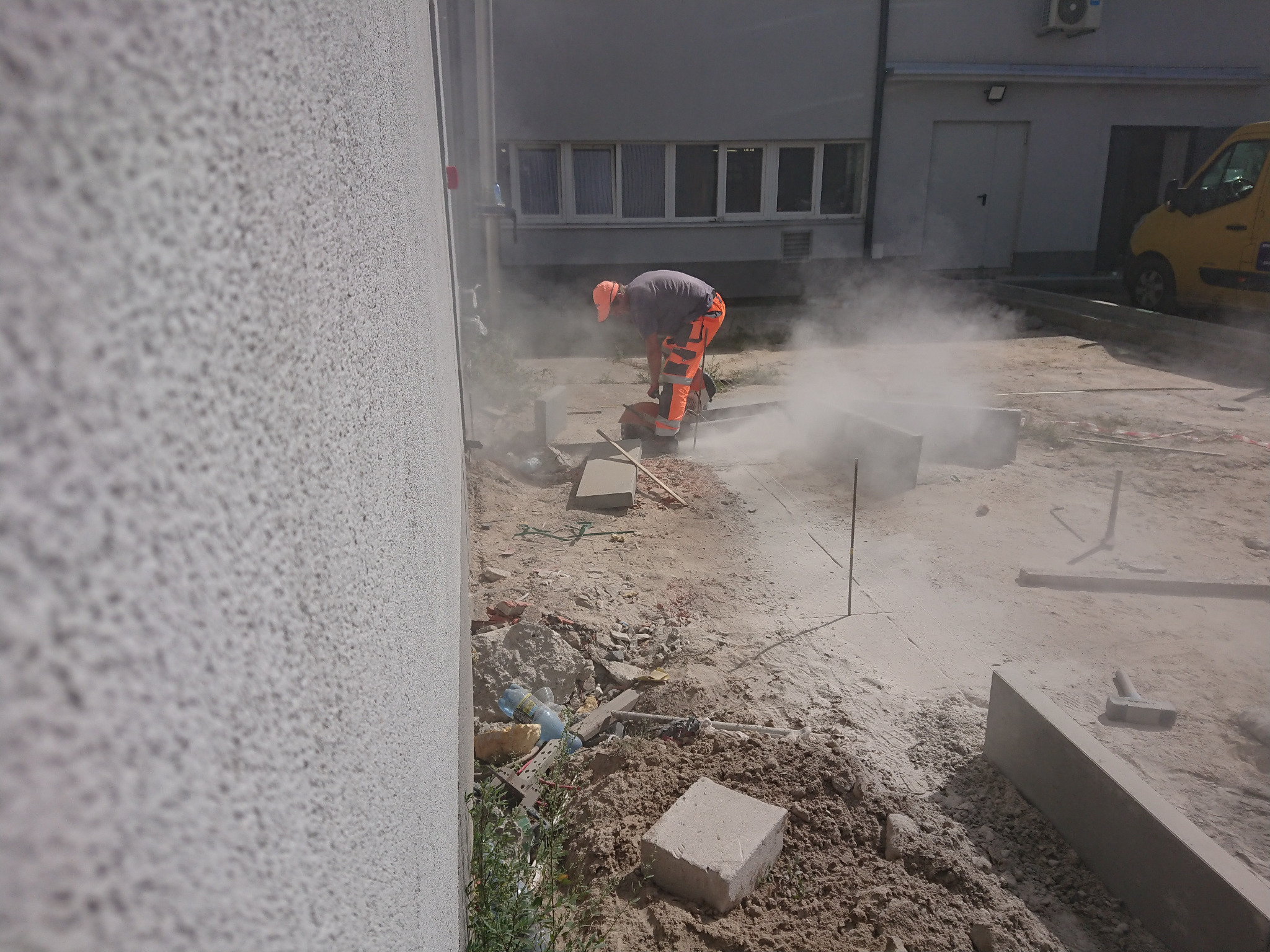 Pracownik w pomarańczowym kombinezonie tnie kostkę brukową za pomocą przecinarki, unoszący się pył, budynek w tle, częściowo widoczny żółty samochód dostawczy.