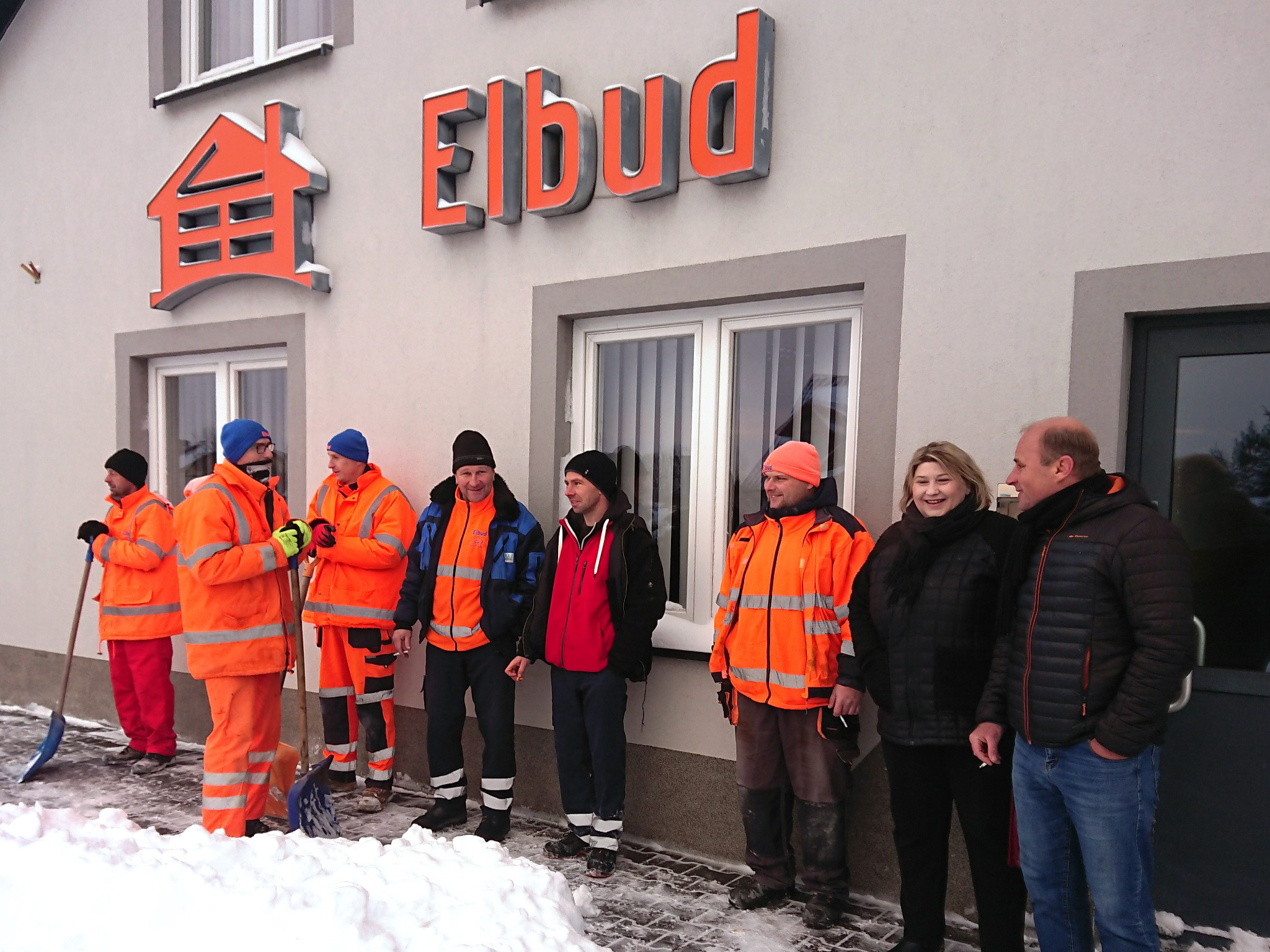 Grupa pracowników firmy budowlanej Elbud z Terespola w strojach roboczych, stojąca przed budynkiem firmy, z łopatami do odśnieżania w tle. Na elewacji widoczne logo firmy.