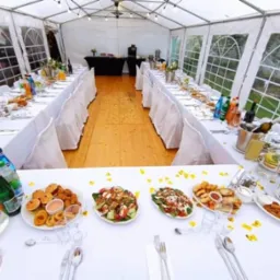Uroczysty st&oacute;ł z białym obrusem i dekoracjami kwiatowymi wewnątrz białego namiotu z oknami, przygotowany na przyjęcie z r&oacute;żnorodnymi potrawami i napojami.