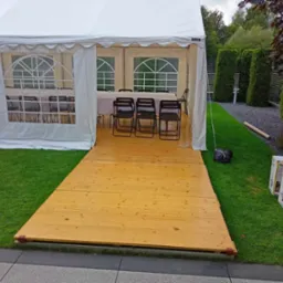 Biały namiot imprezowy z drewnianą podłogą, ustawiony na trawie, widoczne krzesła i stół wewnątrz, paczki z nagrzewnicami w tle.