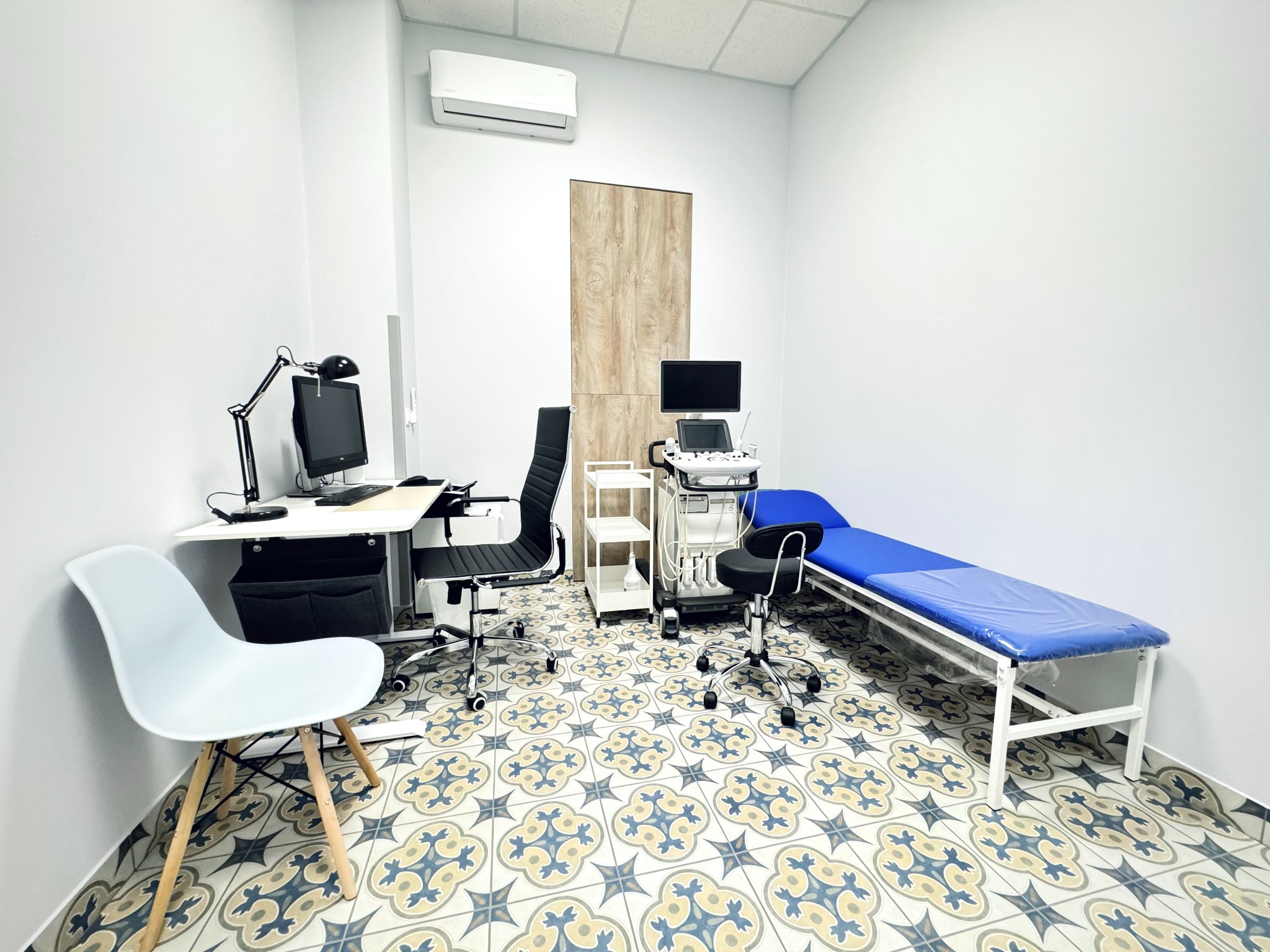 Jasne wnętrze gabinetu lekarskiego z niebieskim leżankiem, aparaturą diagnostyczną na kółkach, biurkiem z komputerem i krzesłami na wzorzystej podłodze.