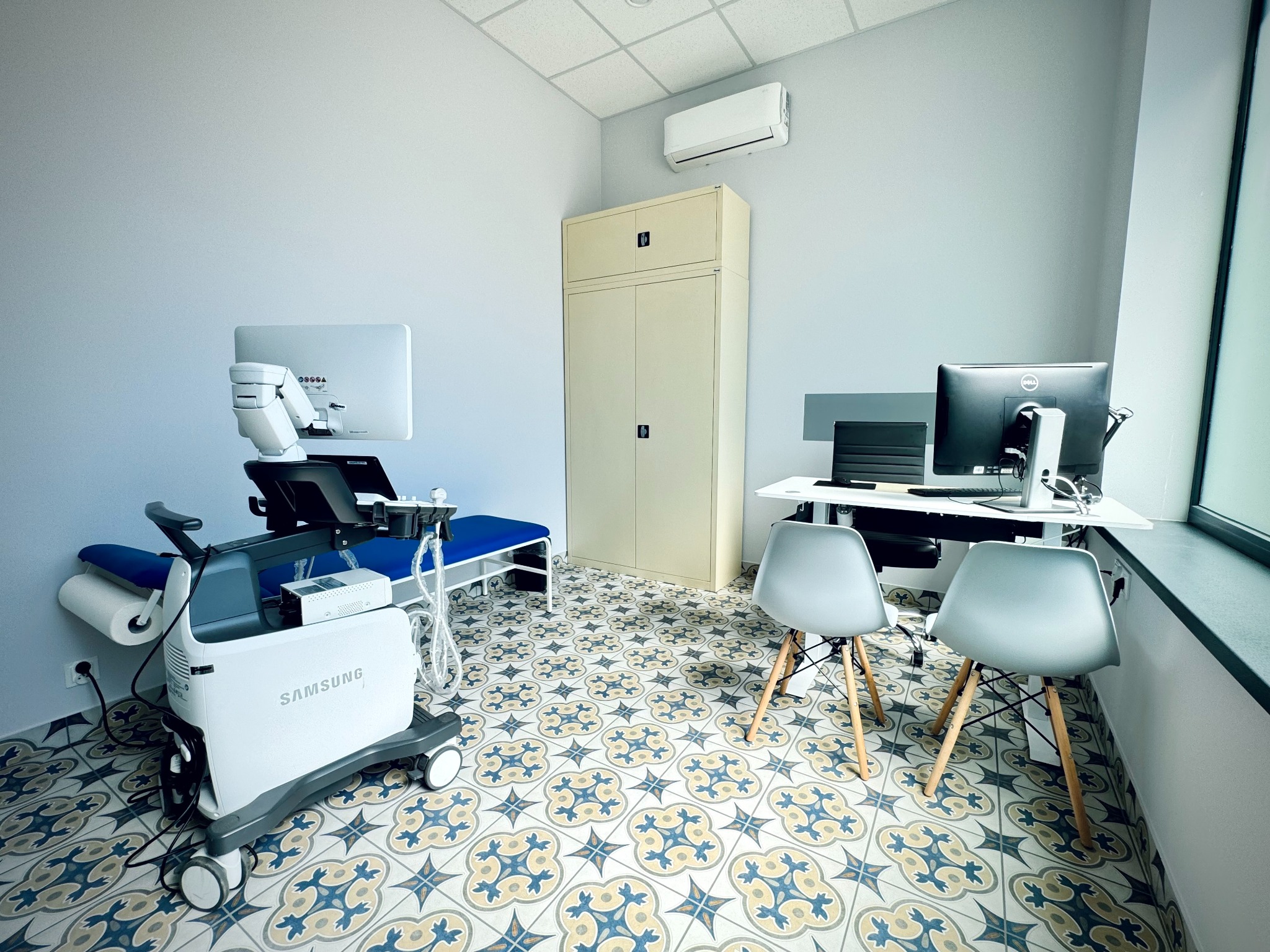 Jasny gabinet lekarski z aparaturą USG marki Samsung, biurkiem z komputerem, szafą i dwoma krzesłami na tle wzorzystej podłogi w stylu retro.