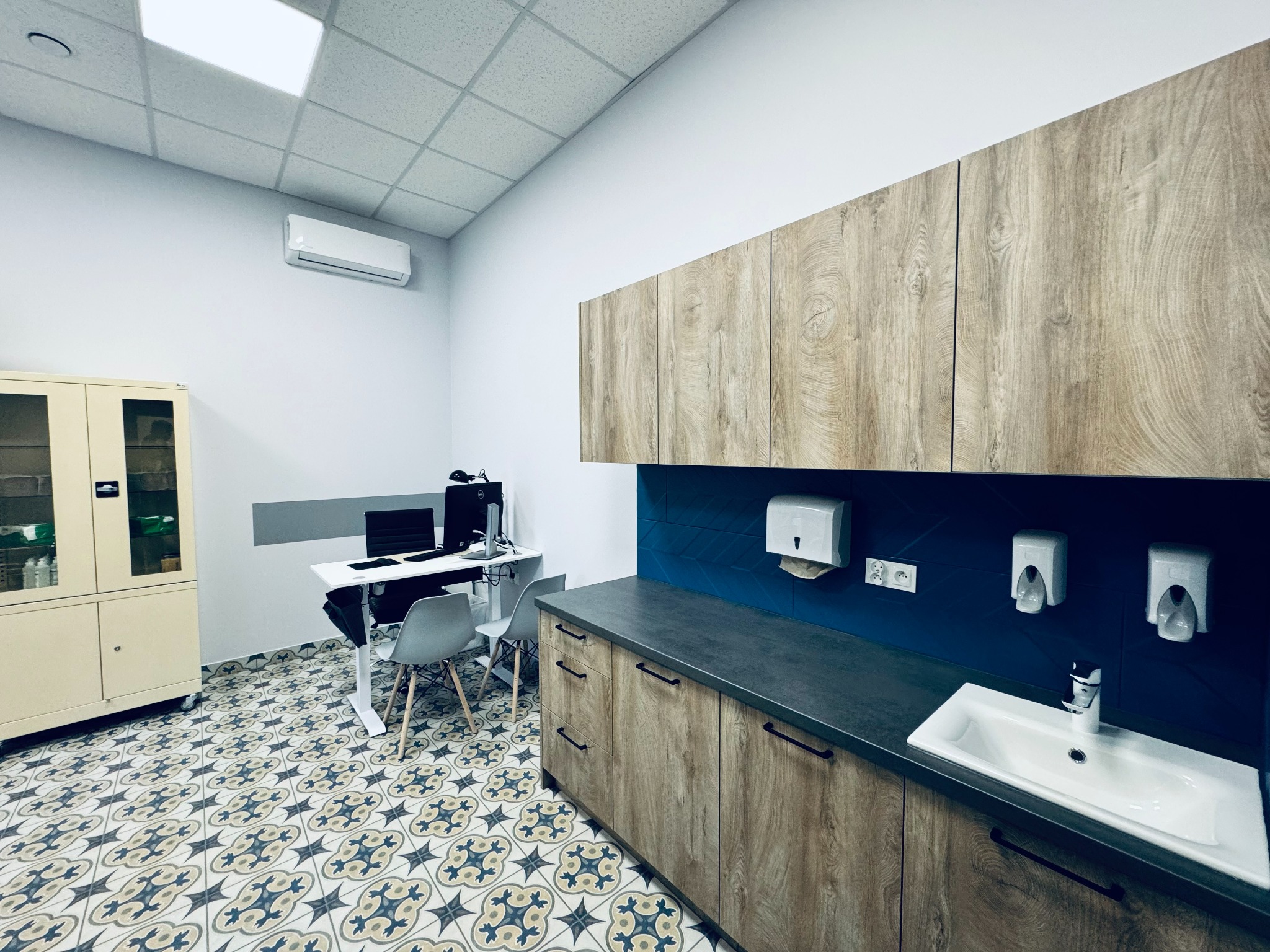 Wnętrze gabinetu lekarskiego z umywalką, szafkami z drewnopodobnym fornirem, wzorzystą podłogą, biurkiem z komputerem i szafką medyczną.