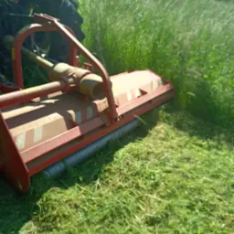 Czerwona kosiarka traktorowa w trakcie koszenia wysokiej, zielonej trawy na polu, widoczne świeżo skoszone źdźbła.