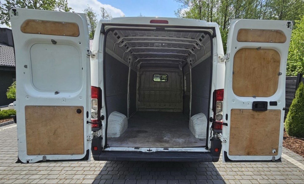 Wnętrze pustej, białej furgonetki z otwartymi tylnymi drzwiami, ukazujące przestrzeń ładunkową wyłożoną szarą wykładziną i drewnianymi panelami na drzwiach. Widok z zewnątrz na kostkę brukową...