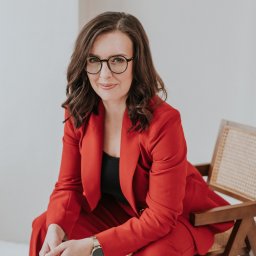 Studio 22 Marta Filipczyk Fotografia - Portret kobiety w czerwonym garniturze, siedzącej na krześle z wiklinowym oparciem, z delikatnym uśmiechem i okularami na tle jasnego, minimalistycznego wnętrza.