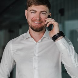 Studio 22 Marta Filipczyk Fotografia - Uśmiechnięty mężczyzna w białej koszuli rozmawia przez telefon, nosi czarny zegarek. Portret w neutralnym wnętrzu, z bliska.