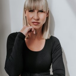 Studio 22 Marta Filipczyk Fotografia - Portret kobiety z blond włosami i grzywką, ubrana w czarną bluzkę z długim rękawem, pozująca na tle jasnej ściany z cieniem. Dłoń podparta pod brodą.