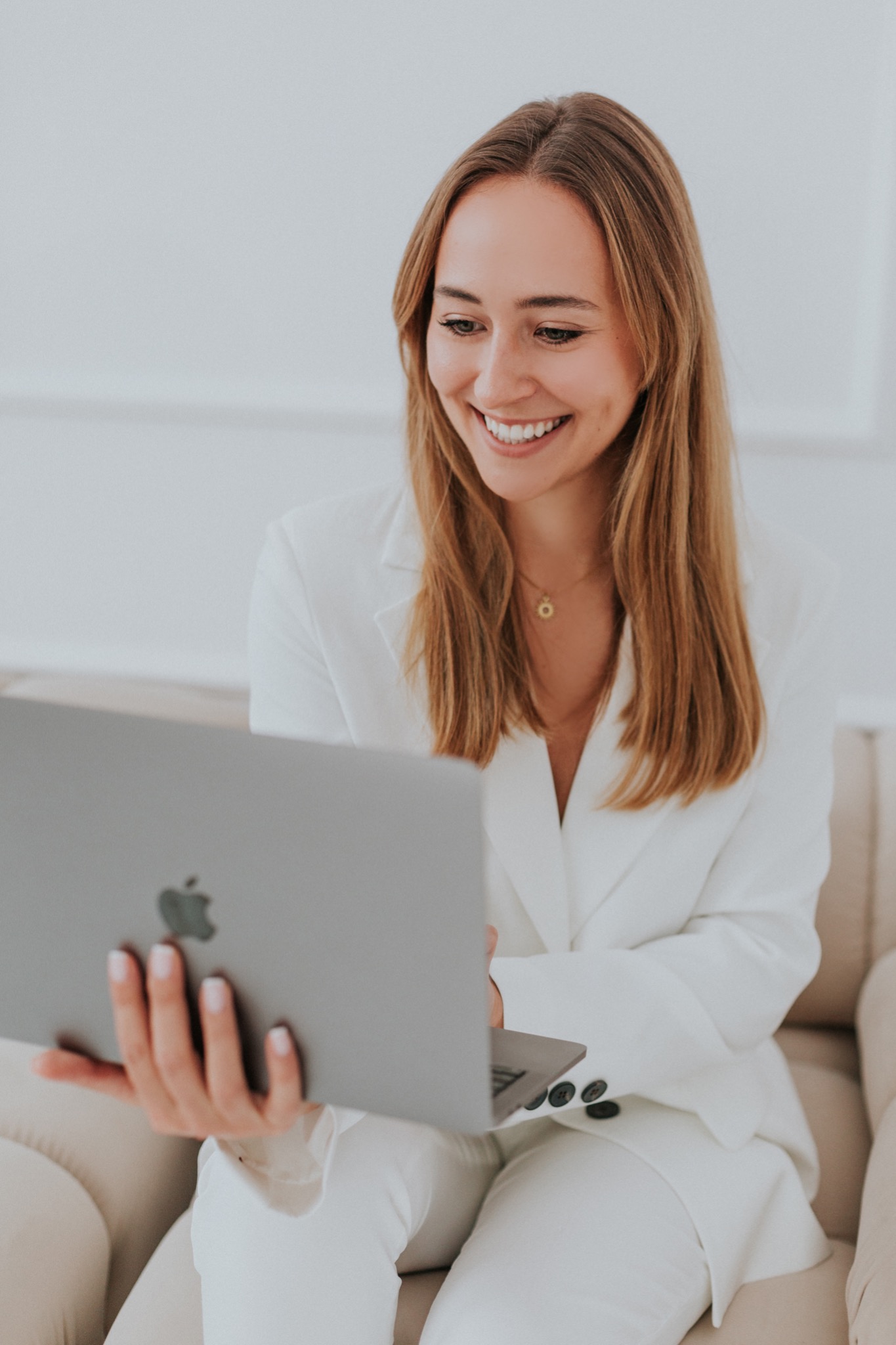 Uśmiechnięta kobieta w białym garniturze siedzi na beżowej kanapie i patrzy w ekran laptopa Apple, trzymanego w dłoniach.