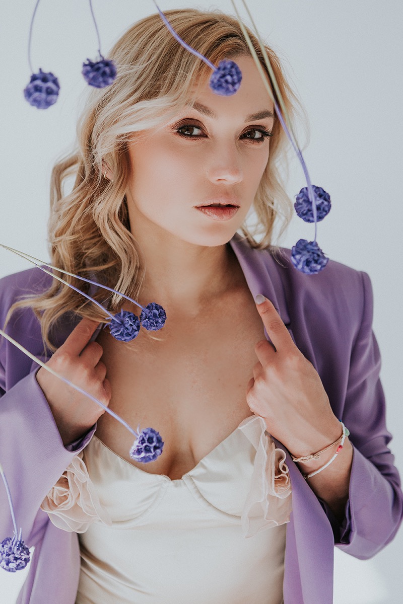 Portret kobiety w fioletowym żakiecie i jasnej bieliźnie, z dekoracjami z fioletowych kwiatów na cienkich łodygach.