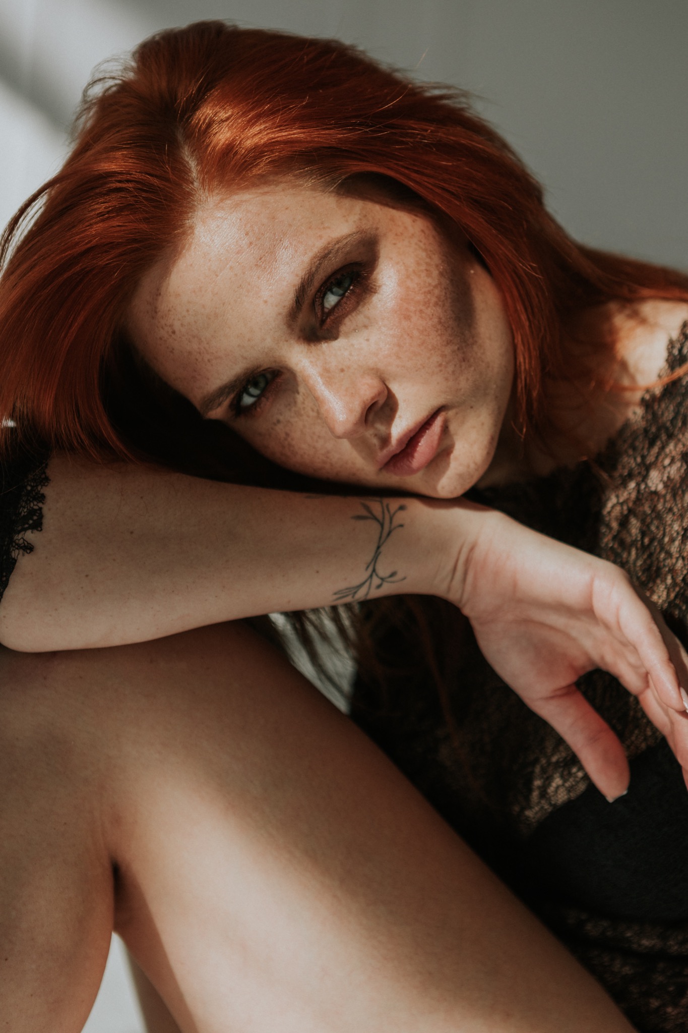Portret rudowłosej kobiety z piegami, opierającej głowę na ramieniu z tatuażem gałązki, w koronkowym topie.