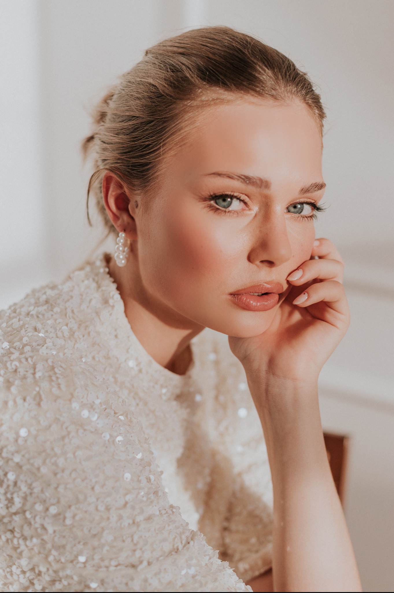 Portret kobiety z delikatnym makijażem, w cekinowej bluzce i perłowych kolczykach, opierającej twarz na dłoni z minimalistycznym manicure.