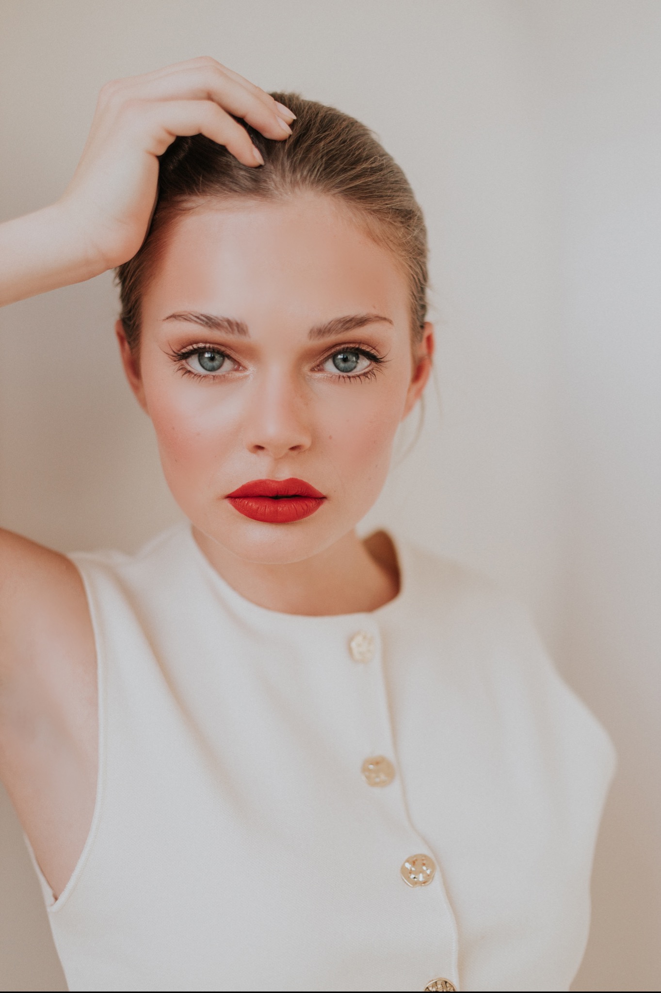 Portret kobiety z intensywnym makijażem, w kremowej sukience z ozdobnymi guzikami, ręka uniesiona do góry. Uwagę przyciągają wyraziste, czerwone usta.