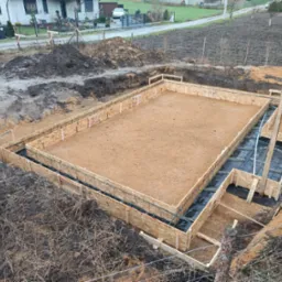 Przygotowanie terenu pod wylewkę fundamentów domu jednorodzinnego, widoczny szalunek z płyty OSB, zbrojenie i izolacja przeciwwilgociowa, ziemia wokół wykopu.