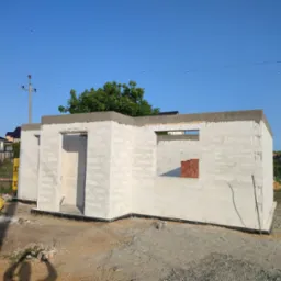 Wznoszona ściana domu z betonu komórkowego z widocznymi otworami okiennymi i pojedynczymi czerwonymi cegłami w jednym z nich, na tle błękitnego nieba i zieleni drzew.