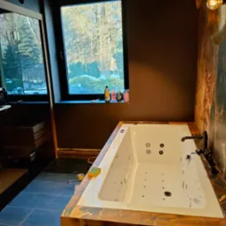 Nowoczesna łazienka z wanną z hydromasażem obudowaną marmurem, ciemne ściany, okno z widokiem na ogród i designerskie oświetlenie ścienne.