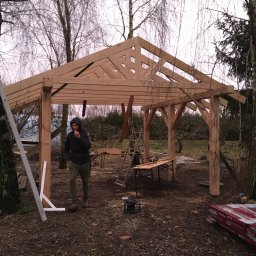 Serhii Haiduchyk - Konstrukcja drewnianej altany w trakcie budowy; widoczny szkielet dachu, słupy nośne, mężczyzna w kurtce i narzędzia na ziemi, drzewa w tle.