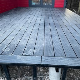 Serhii Haiduchyk - Szary taras kompozytowy z widoczną konstrukcją wsporczą na betonowych słupach, przylegający do czerwonej ściany budynku z szarymi drzwiami balkonowymi.