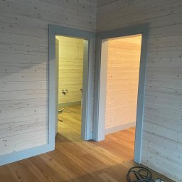 Serhii Haiduchyk - Wnętrze w trakcie wykańczania: ściany wyłożone jasnym drewnem, dwa wejścia z szarymi ościeżnicami, widoczne narzędzia na podłodze z paneli.