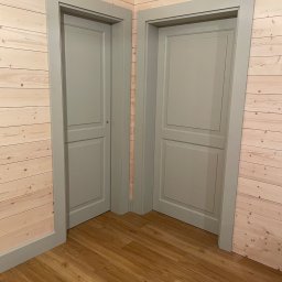 Serhii Haiduchyk - Dwie szare, panelowe drzwi wewnętrzne z ościeżnicami, ustawione pod kątem w rogu pokoju wyłożonego jasnymi panelami drewnianymi, z podłogą z paneli imitujących drewno.