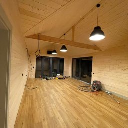 Serhii Haiduchyk - Wnętrze w trakcie wykańczania, ściany i sufit wyłożone jasnym drewnem, na podłodze panele imitujące drewno, widoczne niedokończone instalacje elektryczne i narzędzia pracy.