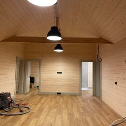 Serhii Haiduchyk - Wnętrze wykończone boazerią w jasnym odcieniu drewna, z trzema czarnymi lampami wiszącymi, widoczne drzwi i odkurzacz przemysłowy na podłodze imitującej drewno.