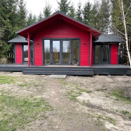 Serhii Haiduchyk - Czerwony domek letniskowy z grafitowym dachem i tarasem na tle lasu, widok z przodu, na ziemi widoczna trawa i piasek.