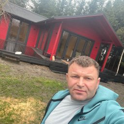 Serhii Haiduchyk - Mężczyzna robi selfie na tle czerwonego domku z czarnym dachem i tarasem, widoczny fragment terenu z błotem i drzewami.