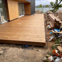Serhii Haiduchyk - Nowoczesny taras z desek drewnianych przylegający do budynku z drewnianą elewacją, widoczne przygotowane deski, ziemia i narzędzia budowlane.