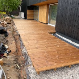 Serhii Haiduchyk - W trakcie budowy tarasu drewnianego przy nowoczesnym domu z czarną elewacją, widoczne deski tarasowe w ciepłym odcieniu brązu, przygotowane podłoże z kamienia i elementy konstrukcyjne w tle.