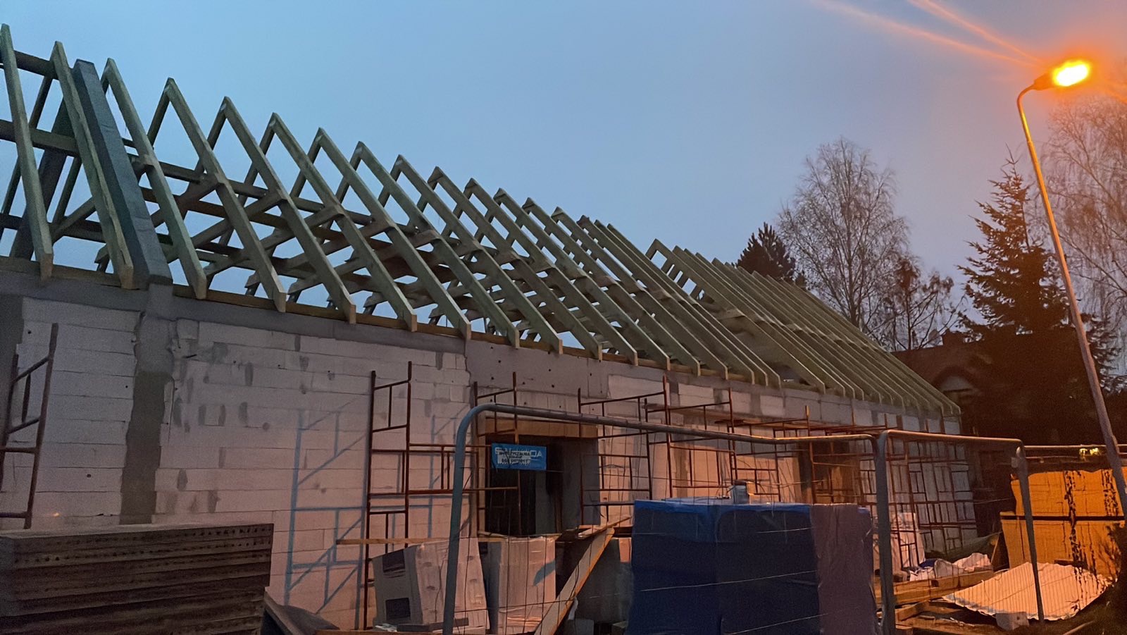 Konstrukcja drewniana więźby dachowej na murowanym budynku w trakcie budowy, widoczne rusztowania i materiały budowlane w tle, oświetlenie uliczne w tle.