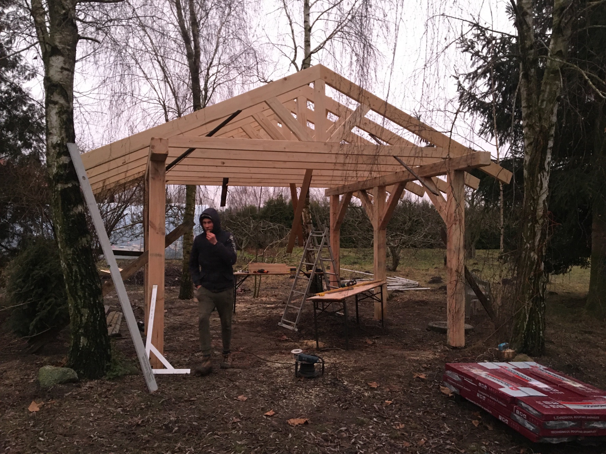 Konstrukcja drewnianej altany w trakcie budowy; widoczny szkielet dachu, słupy nośne, mężczyzna w kurtce i narzędzia na ziemi, drzewa w tle.