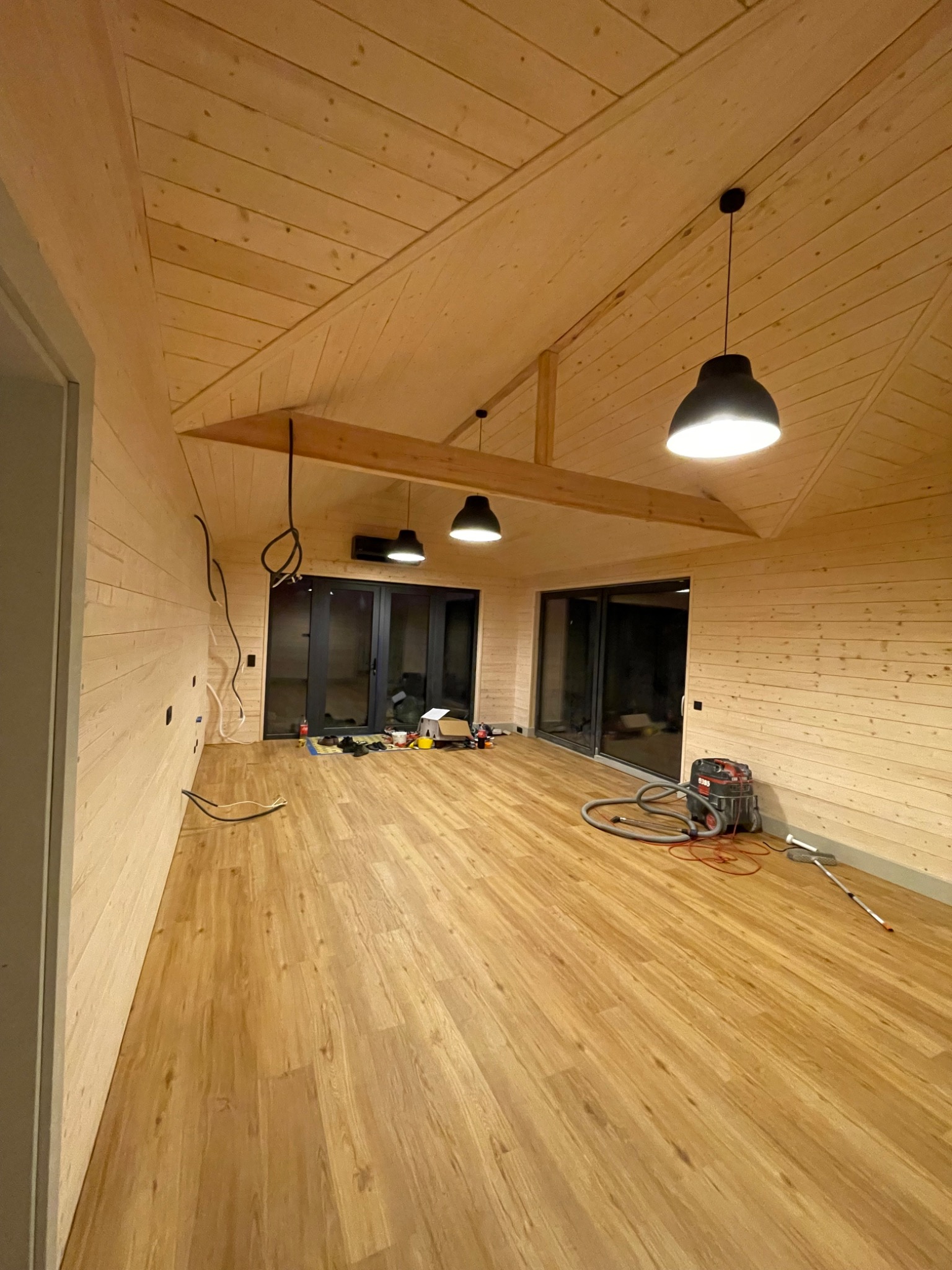 Wnętrze w trakcie wykańczania, ściany i sufit wyłożone jasnym drewnem, na podłodze panele imitujące drewno, widoczne niedokończone instalacje elektryczne i narzędzia pracy.