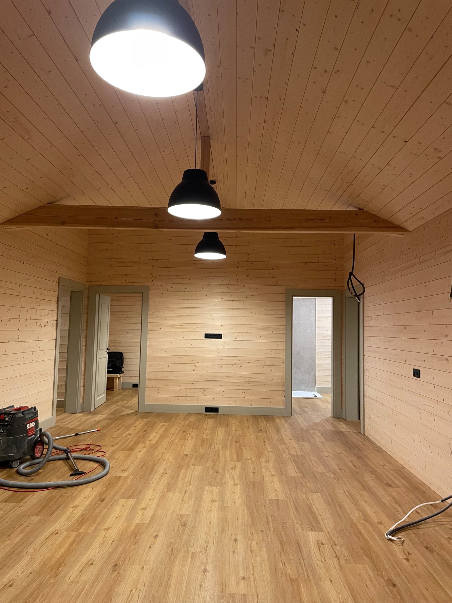 Wnętrze wykończone boazerią w jasnym odcieniu drewna, z trzema czarnymi lampami wiszącymi, widoczne drzwi i odkurzacz przemysłowy na podłodze imitującej drewno.
