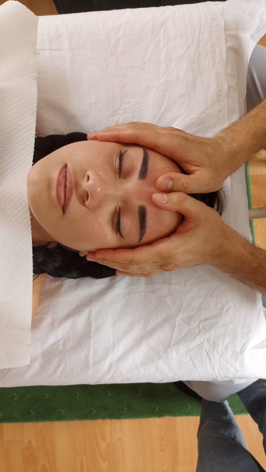 Kobieta z zamkniętymi oczami podczas automasażu twarzy wykonywanego przez masażystę na białym prześcieradle.
