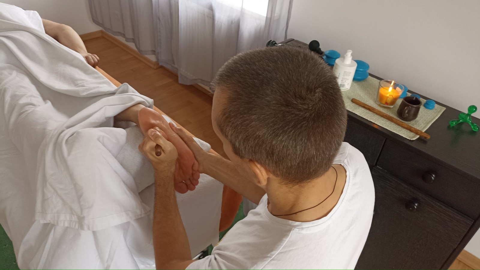 Masażysta wykonuje masaż stóp pacjentowi na leżance w gabinecie, w tle widoczna świeca i akcesoria do masażu na komodzie.