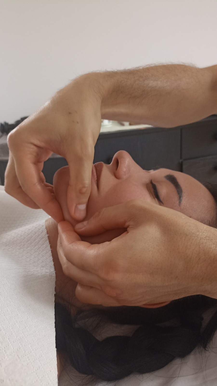 Kobieta leżąca na plecach podczas masażu twarzy, z dłońmi masażysty delikatnie uciskającymi jej policzki i podbródek.