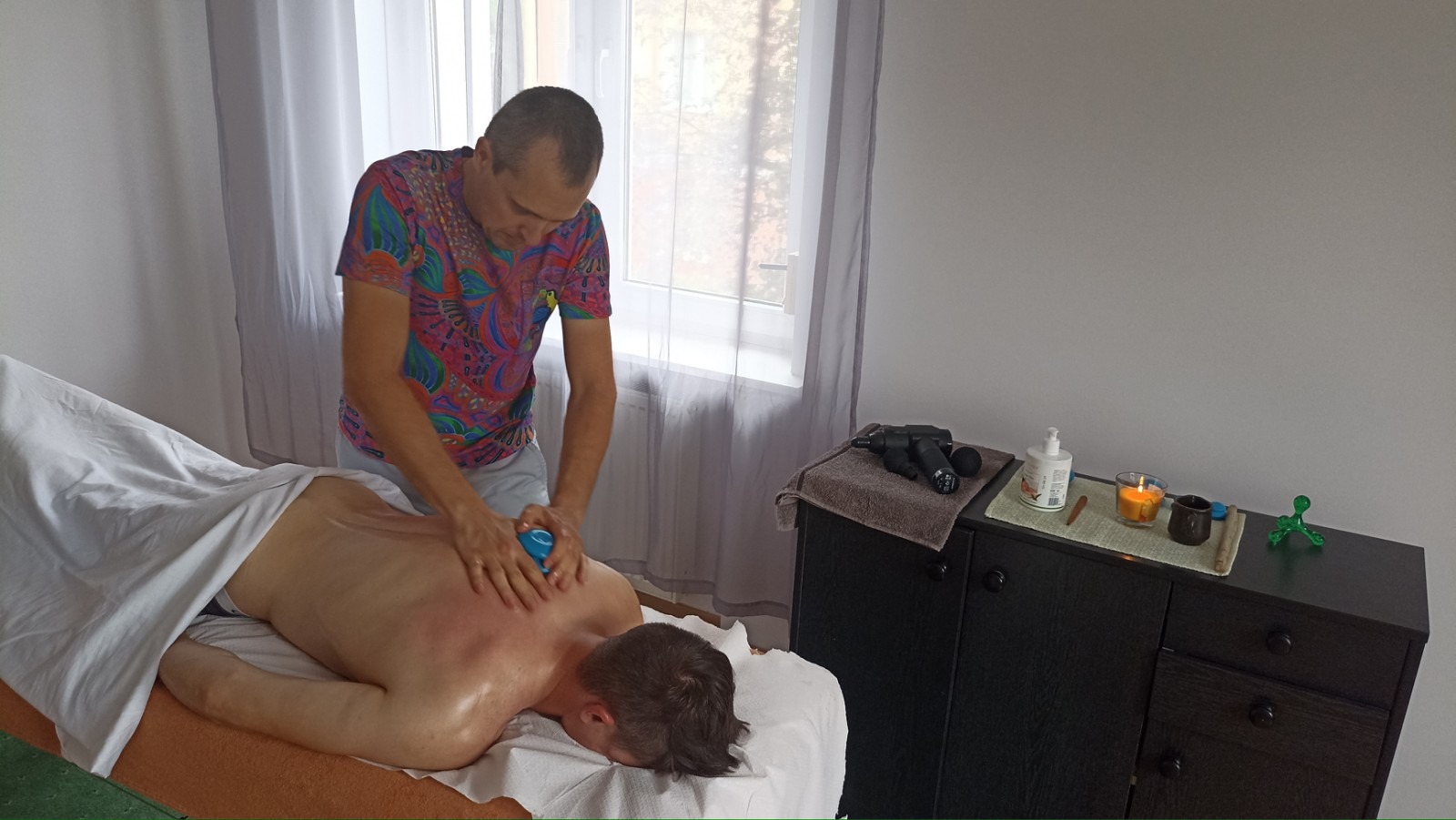 Mężczyzna w kolorowej koszulce wykonuje masaż pleców pacjenta leżącego na stole; widoczne narzędzia do masażu, świeczka i kosmetyki na szafce obok.