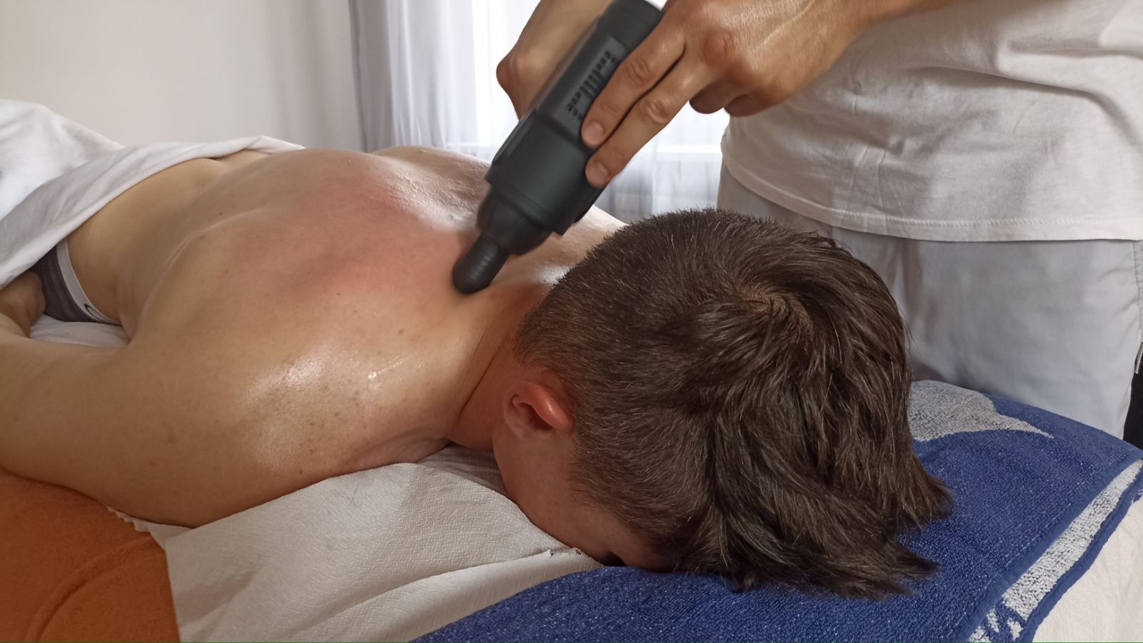 Mężczyzna leży na brzuchu podczas zabiegu z użyciem pistoletu do masażu na górnej części pleców, widoczne zaczerwienienie skóry.
