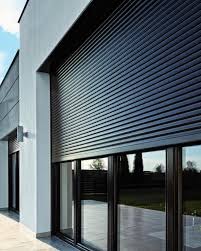 Zamknięta, czarna osłona okienna z lamelami na nowoczesnym budynku z dużymi oknami, widoczne odbicie otoczenia w szybach.