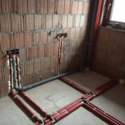 Instalacja hydrauliczna w trakcie montażu w surowym wnętrzu, widoczne czerwone rury grzewcze ułożone na podłodze, szare rury pionowe przy ścianie z cegły, pianka montażowa wokół przyłączy.