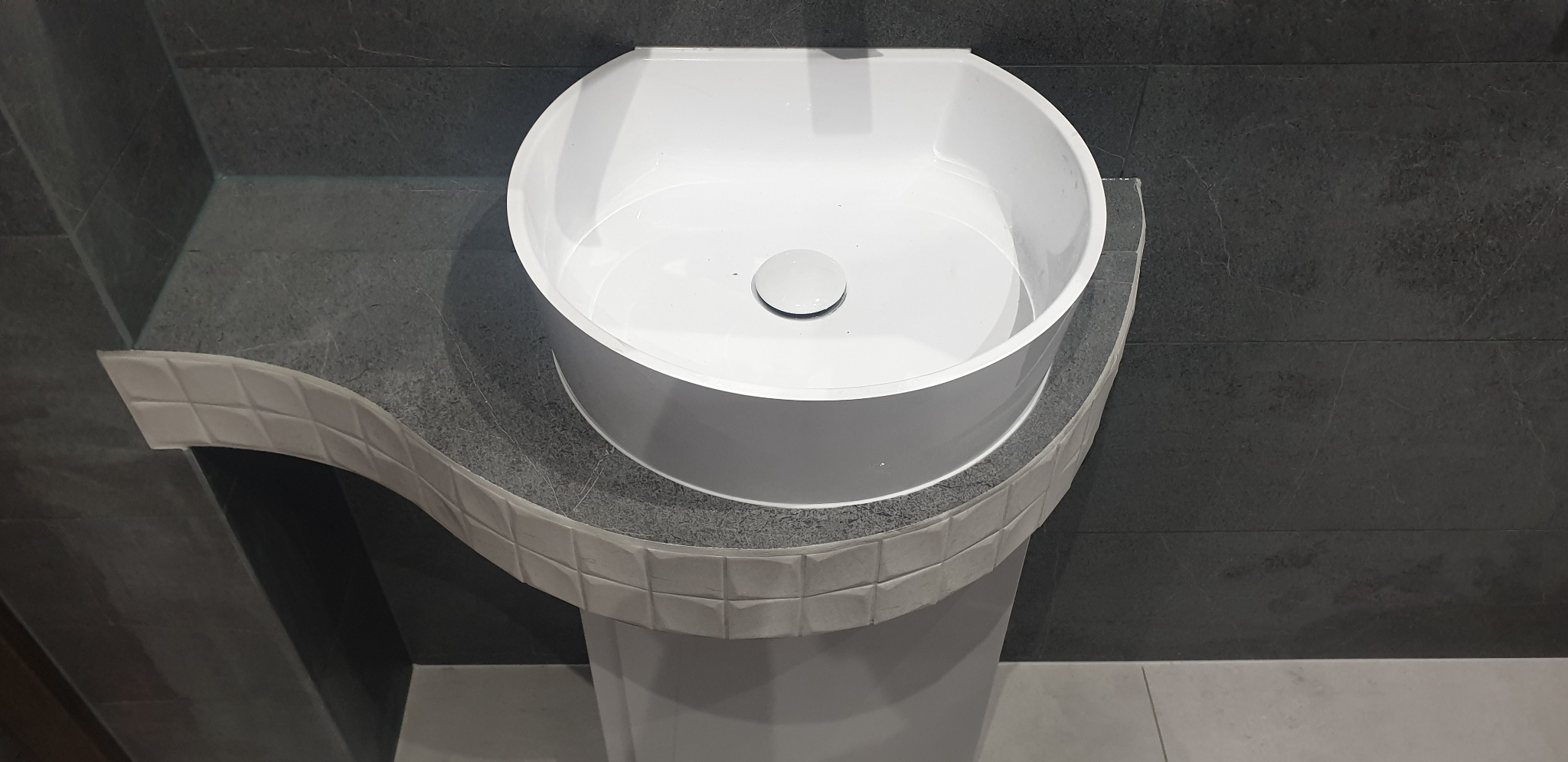 Biała, okrągła umywalka nablatowa z chromowanym korkiem, osadzona na postumencie z szarego kamienia z dekoracyjnym obramowaniem z małych, kwadratowych płytek, na tle ściany wyłożonej ciemnoszarymi...