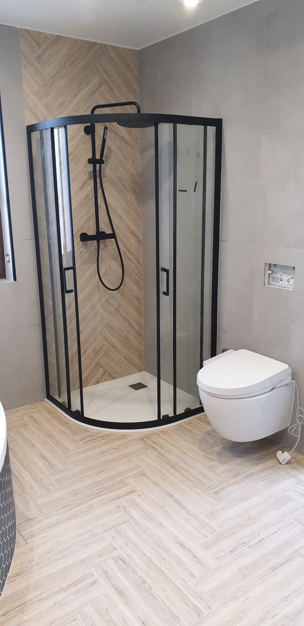Nowoczesna łazienka z kabiną prysznicową w czarnej ramie, toaleta myjąca z podłączeniem elektrycznym i podłoga wyłożona płytkami imitującymi drewno ułożonymi w jodełkę.