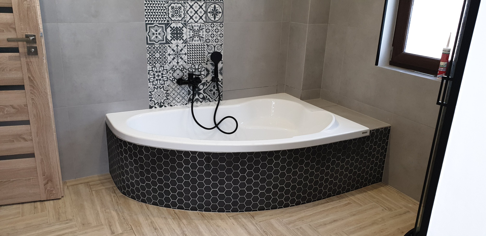 Narożna wanna obudowana czarną mozaiką heksagonalną, z czarną armaturą na tle szarych płytek i dekoracyjnego panelu z wzorzystych kafelek, drewnopodobna podłoga.
