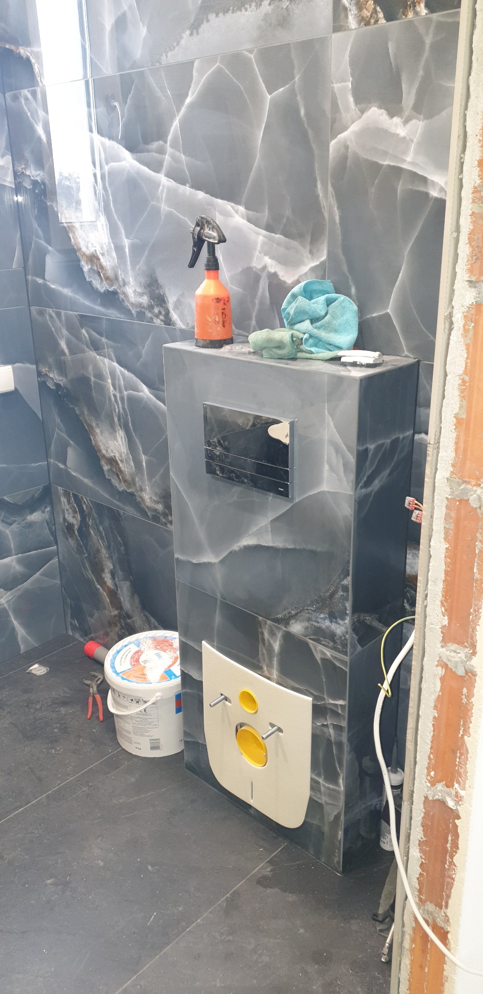 Montaż stelaża podtynkowego WC na tle ściany z płytek imitujących marmur, widoczny spryskiwacz, wiadro z farbą i narzędzia.