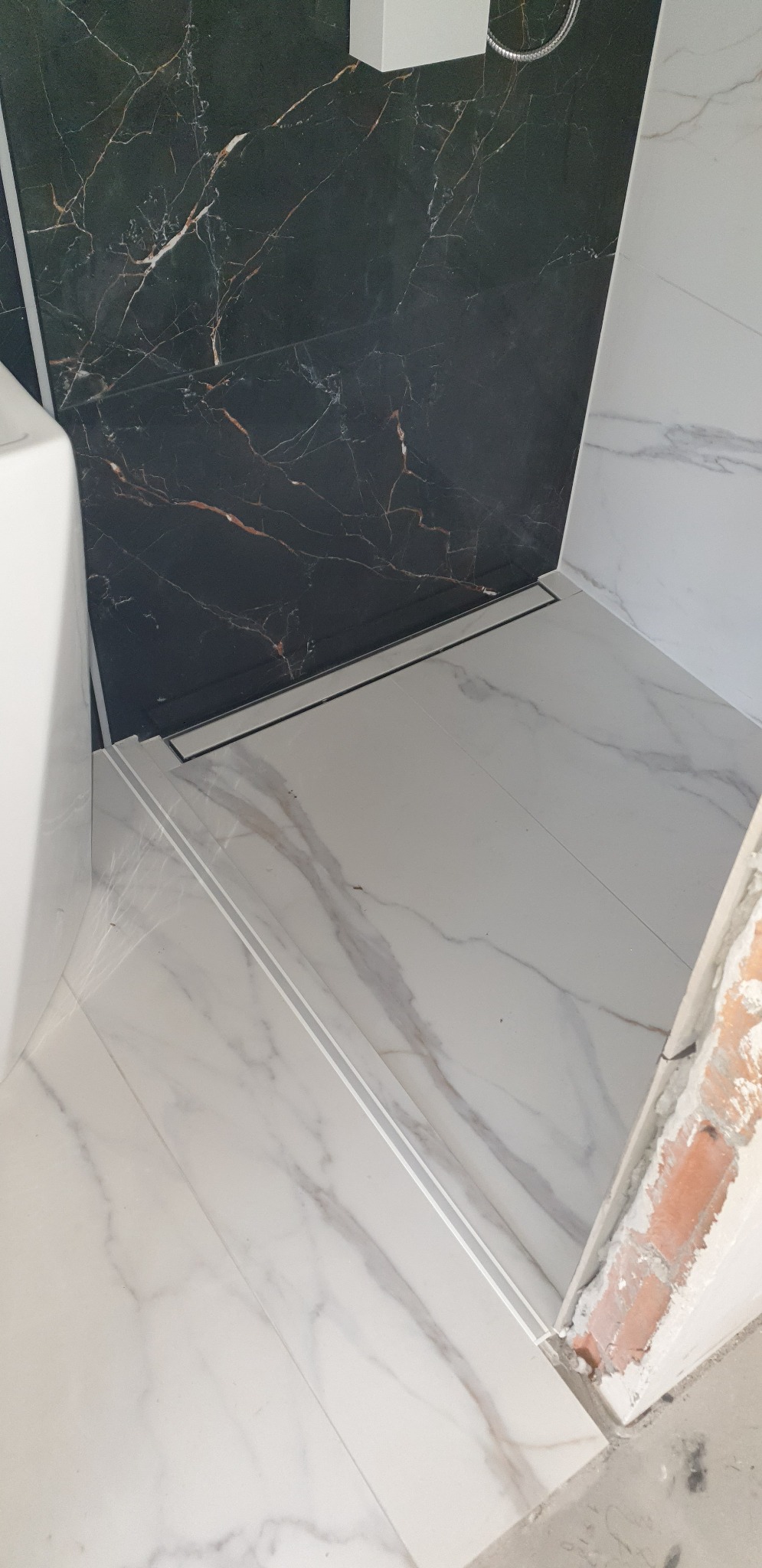 Wykończona łazienka z czarną marmurową ścianą prysznicową, białymi marmurowymi płytkami podłogowymi z odpływem liniowym i niedokończonym wykończeniem drzwi z cegły.