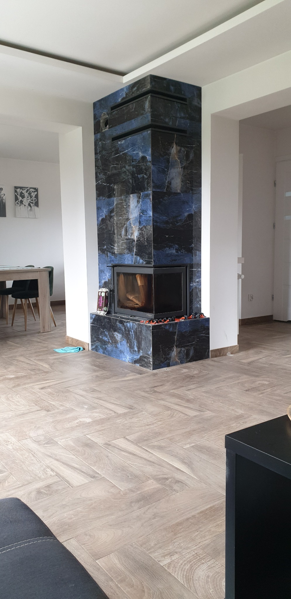 Nowoczesny kominek obłożony ciemnym marmurem z niebieskimi żyłami, widoczny w salonie z drewnopodobną podłogą ułożoną w jodełkę, z fragmentem stołu i krzeseł w tle.